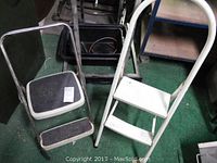Photo showing two folding step stools, one with black steps and one with white steps, placed on green floor.