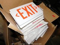 Photo showing a stack of white EXIT signs with orange lettering inside a cardboard box, clearly visible orange text and arrows on each sign.