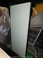 Photo of the oblong folding table in folded position leaning upright showing textured light gray top with black edge trim.