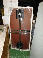 Bottom view of the folding table showing metal support structure and hinges, wrapped in protective plastic.