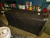 Photo showing the closed large outdoor storage trunk with deep brown plastic and textured woven pattern, next to paint cans and other items.