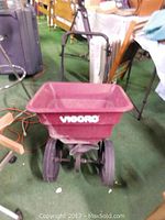 Front view of the Vigoro spreader showing the hopper with brand logo and two rubber tires on a green floor indoors next to other miscellaneous items.