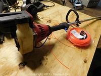 Photo of red and black gas-powered weed trimmer with white fuel tank and loop handle, placed on wood surface beside sealed spool of orange trimmer line.