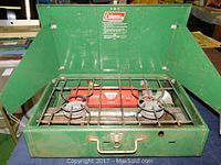 Open view of the green Coleman portable two-burner propane grill showing burners, grill grate, and fold-out wind guards.
