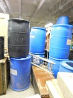Two rain barrels stacked, one black on top of one blue, with additional blue barrels in the background.