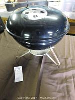Small black Weber charcoal grill with round vented lid and silver tripod base shown from the front
