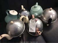 Five assorted vintage tea pots and one extra metallic cover arranged on a surface, showing variety in materials and styles.