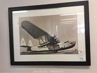 Framed black and white photo of vintage De Havilland seaplane hanging on wall.