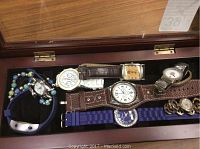 Wooden box with glass lid containing assorted watches with visible detailed faces and bands.