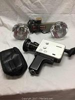 Full view of Nizo S80 movie camera with lens, Bell and Howell dual-head floodlight, and black leather carrying case, all set on white fabric.