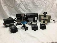Full overview of nine vintage cameras and flashes on white backdrop showing two Polaroid Spirit 600 cameras, Kodak Handle 2 camera, Polaroid Big Swinger 3000 Land Camera, and four camera flashes including Minolta, Canon, and Polaroid models.