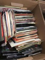 Large box filled with a stack of vinyl 45 RPM records and paper sleeves.