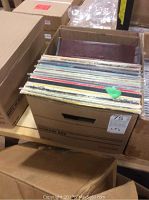 Cardboard box filled with a large number of vinyl LP records visible with different colored spines and some wear on sleeves.