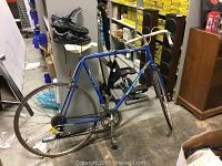 Full view of Velo Sport blue road bicycle in storage room, showing wear and used condition, with Diadora shoes on shelf above.