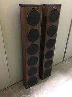 Full view of two tall floor standing speakers with wood veneer and five circular black speaker grilles each, showing size and condition.
