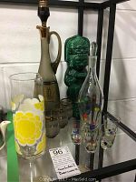 Set of glass barware items including two decanters and multiple small glasses displayed on glass shelving with a green tiki ceramic figure in background.