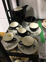 Photo showing 5 cups & saucers, a tea pot, creamer jug, and a small bowl with black and cream color scheme and leaf designs