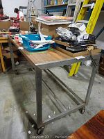 Side view of the rectangular table showing metal frame with wood top surface and items on top, placed on a warehouse floor.