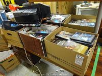Several cardboard boxes filled with assorted DVDs and a boxed IMAX collection set on a table in a warehouse.