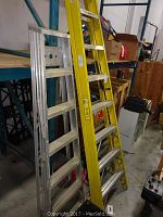 Two aluminum ladders, one silver 90" and one yellow 72", leaning against shelving in storage area.