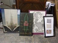 Four framed art prints arranged side by side showing variety in styles and colors. The leftmost print depicts a skyscraper with ships at its base, the next is an Art Deco poster, then a monochrome drawing of a woman, and lastly a colorful abstract print.