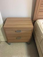 Wooden nightstand shown from the front, two drawers visible with metal handles, standing on short silver legs beside a bed.