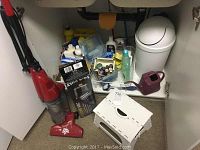 Photo of the Dirt Devil vacuum, white step stool, maroon watering can, and small white garbage can inside a cabinet with other supplies