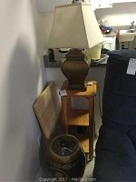 Photo showing table lamp, wooden plant stand, partly visible ceramic barrel with lid, next to a blue couch.