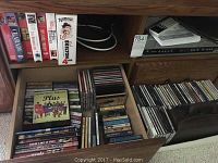 Image showing open wooden cabinet drawers filled with CDs, DVDs, and VHS tapes. Top shelf has VHS tapes and electronic cables. Middle drawer and bottom drawer contain a mixed collection of DVDs and CDs.