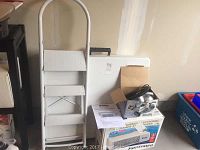 Photo of white metal folding step ladder with three steps, white folding table behind it, Shark hand vacuum box and Kenmore air cleaner box underneath.