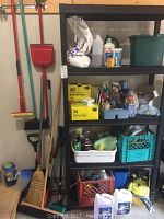 Full view of black plastic shelving unit with various cleaning and maintenance items including brooms, dustpans, shovel, buckets, and storage crates on and around it.