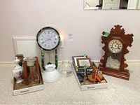 Photo showing several items including carved wooden gingerbread style clock, large mantle or wall clock with green accents, assorted glass and pottery items, and tobacco pipe in boxes on carpeted floor.