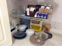 Overall view of various kitchen items including casseroles, cups, trays, storage bins, and tins arranged on a shelf and floor