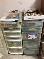 Photo showing two plastic organizers with multiple drawers holding crafting supplies, a clear plastic box of rubber stamps on top of a white organizer, and a cardboard box containing assorted seashells.