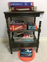 Tri-level side table holding various board games and a plastic container with small items, exercise wrist weights placed beneath the table on the floor.