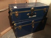 First photo showing both blue steamer trunks stacked in a corner, details of brass hardware and scuffed surfaces visible.