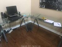 Full view of the L-shaped glass top office desk with metal frame installed in a corner. Computer and some office supplies are on the desk surface.