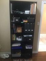 Front view of the black wood bookcase filled with various office and household items only for staging, not included in the lot.