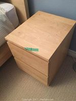 Nightstand positioned next to a bed showing front and side views, top surface has slight wear with a piece of tape attached.