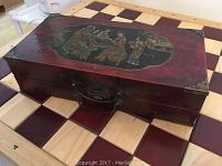 Closed wooden case with red lacquer finish and painted Chinese figure scene on lid, positioned on a checkered surface