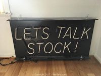 Full view of the "LET'S TALK STOCK!" neon sign mounted on black metal backboard.