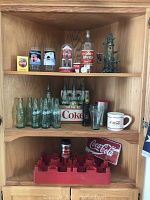 Wooden shelving displaying various vintage Coca-Cola collectible bottles, glasses, mug, metal sign, red plastic crate with bottle, and decorative items.