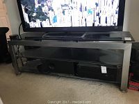 Front view of the glass TV entertainment center showing the metal frame and glass shelves with a TV on top.