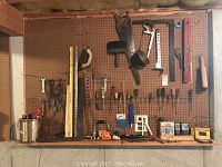 Full view of the wall pegboard with various hand tools including saw, clamps, screwdrivers, funnels and measuring tapes arranged on shelf and hooks.