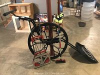 Photo showing Wovel snow shovel with black plastic blade, long handle, and grip, Milwaukee dolly with red accents and black bungee cords, and Mallory snow scraper with black blade, displayed indoors on a concrete floor.