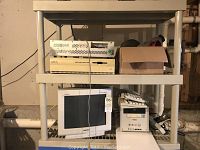Shelf containing various computer components and accessories: top shelf has Shuttle XPC Plus CD Writer and cardboard box; below is beige Opus Personal Mainframe; underneath is white CRT monitor, keyboards and other items.