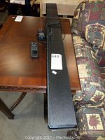 Full view of the RCA sound bar on a wooden table next to a patterned couch, showing the unit with attached power cord.