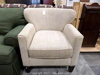 Front view of beige upholstered armchair showing squared arms, cushioned seat, and dark wooden legs, seated next to a green couch.
