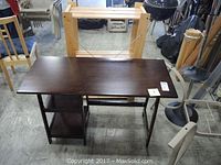 Wooden desk with two shelves, dark finish, visible surface scratches, placed indoors on concrete floor with other furniture around.