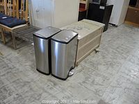 Photo shows two stainless steel metal trash cans next to a weathered beige deck storage bin and metal wire shelves to the left.